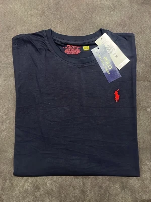 Mörkblå t-shirt från Polo Ralph Lauren - Snygg mörkblå t-shirt från Polo Ralph Lauren med klassisk röd logga broderad på bröstet. T-shirten har rund halsringning och korta ärmar. Tillverkad i mjuk bomull för skön känsla och enkel stil.