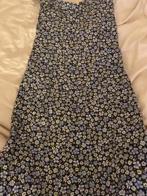 Blommig svart klänning från DIVIDED - Supersöt svart långklänning från DIVIDED i storlek 34/XS med blommigt mönster i blått, vitt och grönt. Klänningen har en rak form. Perfekt för dig som gillar färgglada prints och vill ha en enkel men snygg look. V-ringad framtill och dragkedja på sidan, dessvärre ett litet hål ovanför dragkedjan men det är inget som märks