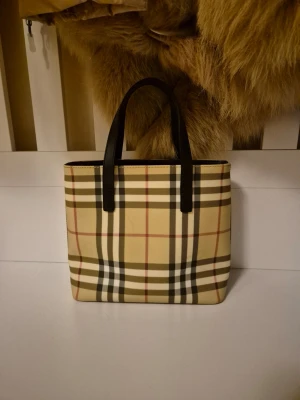 Handväska från Burberry - Säljer en klassisk handväska från Burberry med det ikoniska rutiga mönstret i beige, svart, vitt och rött. Väskan har svarta handtag i skinn och en fyrkantig form . Perfekt för dig som gillar tidlös design och vill sticka ut.