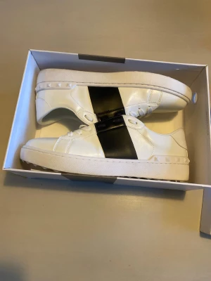 Valentino Garavani vita sneakers - Säljer ett par snygga vita Valentino Garavani sneakers med bred svart rem över sidan. Skorna har platt sula med rutmönster och coola nitar runt hälen. Tillverkade i skinn med snörning och rund tå. Perfekta för dig som gillar stilrena och lyxiga sneakers.