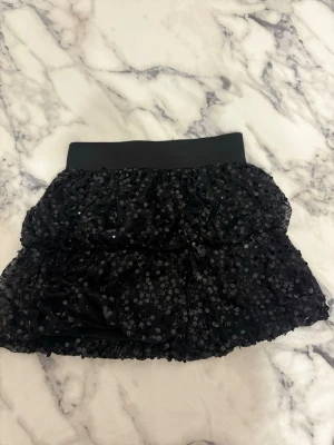 Svart paljettkjol från KappAhl - Säljer en svart kortkjol från KappAhl med glittriga paljetter över hela tyget. Kjolen har ett brett, elastiskt midjeband och volangdetaljer som ger extra fluff. Perfekt för dig som gillar att sticka ut med lite extra bling.