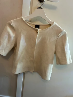 Beige kort kofta från Gina Tricot - Snygg beige kofta från Gina Tricot i kort modell med rund hals och en guldfärgad knapp framtill. Koftan har korta ärmar 