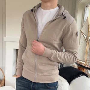 Beige cardigan jacka från Colmar Originals - Snygg beige fleecejacka från Colmar Originals med huva i ljusare ton och dragkedja framtill. Jackan har ribbade muddar, två fickor och en broderad logga på ärmen. Perfekt för chill dagar och har en clean, modern look.