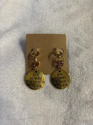 Guldiga örhängen med budskap - Snygga örhängen i guldfärgad metall med runda hängen där det står 'She believed she could so she did'. Perfekt accessoar för dig som gillar inspirerande smycken och vill sticka ut lite extra.