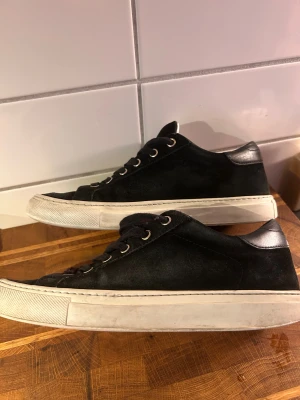 Svarta mockasneakers från Sandays - Svarta sneakers från Sandays med ovandel i mocka och detaljer i svart skinn vid hälen. Skorna har snörning med metallöglor och en vit platt sula. Snygg och stilren design med rund tå och diskret logga i guld på plösen. Litet hål under skon men går inte in i skon. Nypris 2199:-