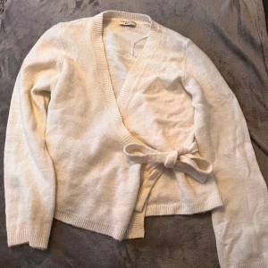 omlottkofta Gina Tricot  - Mysig vit omlottkofta från Gina Tricot i storlek XL. Koftan har långärmade ärmar, ribbade muddar och knytband i midjan. Tillverkad i mjukt stickat material som känns skönt mot huden. Perfekt för att ge din outfit en chill och fräsch vibe.