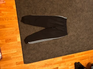 Svarta Adidas mjukisbyxor med vita ränder - Svarta mjukisbyxor från Adidas med klassiska vita ränder längs sidorna och Adidas-logga på vänster lår. Byxorna har elastisk midja med snörning och muddar vid bensluten. Perfekta för chill eller träning.
