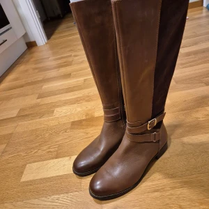 Helt nya bruna höga stövlar med spänne, strl 39 - Snygga helt nya höga stövlar i brunt skinn och mocka med rund tå och platt sula. Stövlarna har guldfärgad dragkedja på sidan och dekorativt spänne runt ankeln. Perfekt till höst och vinter för en stilren look.