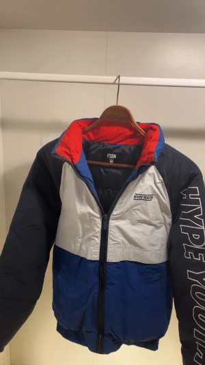Färgglad pufferjacka från FSBN XS - Säljer en pufferjacka från FSBN i storlek XS. Jackan har cool färgblockdesign i blått, vitt och rött, med 'HYPE YOUTH'-tryck på ena ärmen. Dragkedja framtill och hög krage. Perfekt för dig som vill sticka ut med streetstyle-vibe.