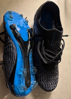 Under Armour Magnetico blå fotbollsskor - Säljer ett par Under Armour Magnetico fotbollsskor i svart och blått vågigt mönster på ovansidan. Skorna har snörning, platt sula med dobbar och stor UA-logga på sidan. Ovandelen är i syntetmaterial för bra passform och grepp på planen. Helt oanvända, köpta nyligen men för små. Passar 41-42. Nypris 3100 cirka säljer för 2000kr.