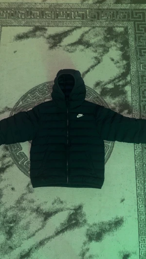 Nike jacka  - Nike pufferjacka i svart med huva och diskret logga på bröstet. Jackan har horisontella quiltade sektioner, två sidofickor och hel dragkedja framtill. Perfekt för kalla dagar och har en clean, sportig vibe. Passformen är normal och jackan är långärmad.