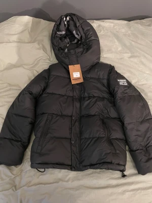 Svart pufferjacka från Burberry - Svart pufferjacka från Burberry med huva och dragkedja, samt knappar framtill. Jackan har Burberry London England-tryck på ärmen och klassiskt rutigt foder i huvan. Två sidofickor med dragkedja och justerbar nederkant. Jackan har även antagningsbara ärmar så den kan bli en väst också. Perfekt för kalla dagar. Jackan är liten i storleken! Pris går att diskutera vid snabb affär