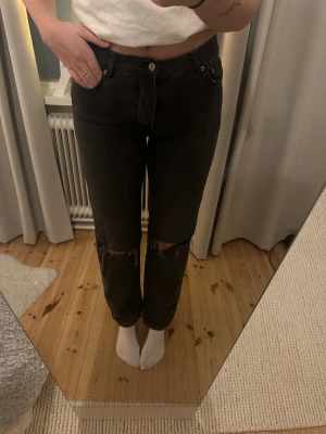 Svarta ripped jeans från Junkyard - Säljer ett par svarta Nevermind Jeans från Junkyard med snygga slitningar över knäna. Modellen har hög midja, raka ben och klassisk femficksdesign. Jeansen är tillverkade i jeansmaterial och har knappgylf framtill.