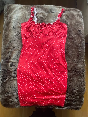 Röd kortklänning med hjärtan och volang - Supersöt röd kortklänning med vita hjärtan, volangkant och knytband framtill. Axelbanden har volangdetaljer i rött och vitt som ger en gullig vibe. Materialet är mjukt och stretchigt, perfekt för dig som gillar att sticka ut med färg och mönster.