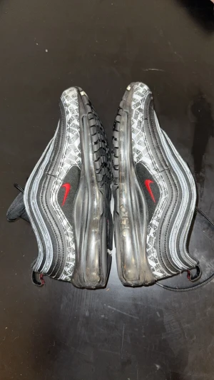 Nike Air Max 97 svart och töd - Nike Air Max 97 sneakers i svart och röd med röda swoosh-detaljer. Ovandelen har ett mönster med Nike-loggor och reflekterande linjer. Sulan är genomskinlig och har en synlig Air-enhet. Skorna har snörning och rund tå, tillverkade i syntet och textil.
