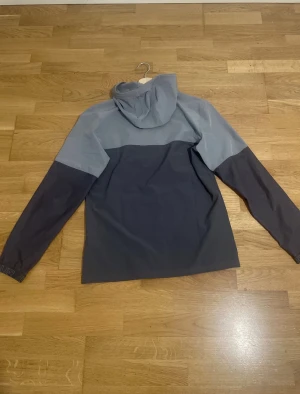 Grå vindjacka från Under Armour - Snygg tvåfärgad vindjacka från Under Armour i ljusgrått och mörkgrått. Jackan har huva, dragkedja framtill och praktiska fickor. Perfekt för dig som vill ha en sportig look och gillar funktionella detaljer. Under Armour-logga på bröstet för extra stilpoäng Går inte ner mer än 50kr!.
