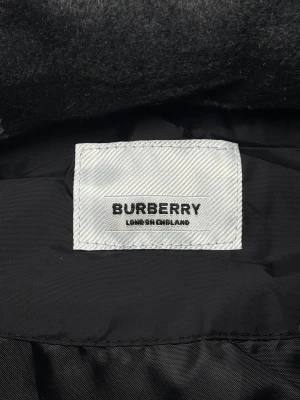 Svart pufferväst från Burberry - Svart pufferväst från Burberry med avtagbar huva i grått och vitt rutmönster. Västen har två fickor med dragkedja och knappar framtill. Insidan är fodrad med slätt material och har snygga Burberry-loggor. Perfekt för dig som vill ha en clean och stilren look.