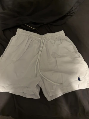 Vita badshorts Polo Ralph Lauren XL - Snygga vita badshorts från Polo Ralph Lauren i storlek XL. Klassisk design med elastisk midja och snörning, sidofickor och bakficka med lock. Broderad marinblå logga framtill. Perfekta för stranden eller poolen.