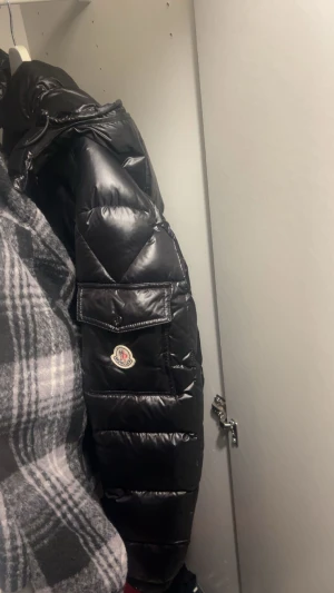Svart pufferjacka från Moncler - Säljer en svart pufferjacka från Moncler med glansig finish och klassisk logga på ärmen. Jackan har praktiska fickor med lock och dragkedja framtill. Perfekt för dig som vill ha en snygg och varm vinterjacka med exklusiv känsla.