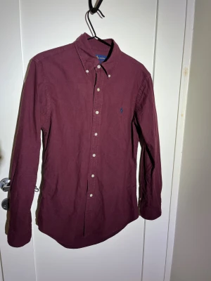 Vinröd skjorta från Ralph Lauren - Vinröd skjorta från Ralph Lauren med klassisk button-down krage och broderad logga på bröstet. Skjortan har långa ärmar, vita knappar och är tillverkad i mjuk bomull. Perfekt för dig som gillar stilrena och tidlösa plagg.
