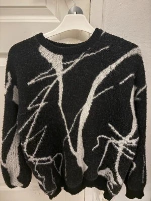 Svart stickad tröja med vit abstrakt design - Cool svart stickad tröja med vit abstrakt mönster över hela. Tröjan har rund hals och långa ärmar, perfekt för dig som gillar en unik och edgy stil. Materialet är mjukt och ger en skön känsla, passar bra till jeans eller cargo pants.