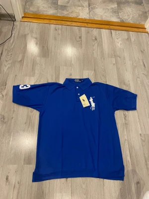 Blå Polo Ralph Lauren pikétröja XL - Blå pikétröja från Polo Ralph Lauren i storlek XL. Klassisk krage, två knappar och stor vit broderad polospelare på bröstet. Siffra 3 på ärmen. Tillverkad i mjuk bomull, perfekt för en clean och sportig look.