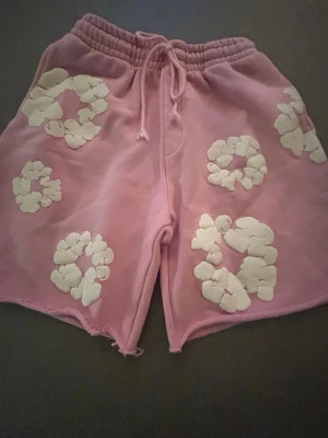 Rosa shorts från Denim Tears med blommor - Säljer ett par rosa denim shorts  med vita blommiga mönster över hela. De har elastisk midja med snörning och råa kanter nertill för en avslappnad vibe. Mycket bra för sommaren. Dom är i storleken S. Priser går att diskutera.