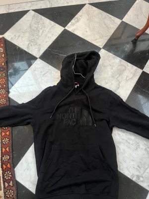 Svart hoodie från The North Face - Svart hoodie från The North Face med stor logga i svart på bröstet. Tröjan har huva med snörning, magficka och långa ärmar. Mjuk och skön bomullsmix, perfekt för chill eller streetwear. Klassisk och enkel design som funkar till allt.