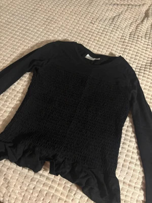 Svart långärmad topp med volang Gina Tricot - Svart långärmad topp från Gina Tricot med smockad front och volangkant nedtill. Rund halsringning och mjukt, stretchigt material som sitter skönt. Perfekt för dig som gillar stilrena plagg med en twist. Passar dig som vill ha något enkelt men ändå med coola detaljer.