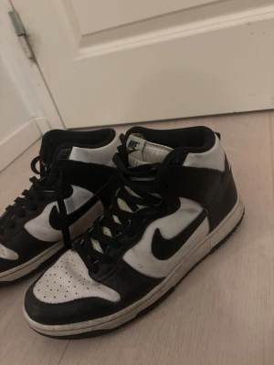 Nike Dunk High svart/vit sneakers - Säljer ett par Nike Dunk High sneakers i svart och vitt skinn med klassisk swoosh på sidorna. Skorna har svarta snören, perforerad tå och hög siluett som ger en riktigt clean och tidlös look. Perfekta för dig som gillar streetstyle och vill sticka ut med en ikonisk modell.