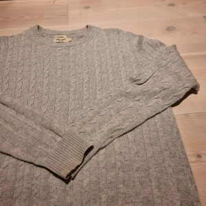 Grå kabelstickad tröja East West - Säljer en grå kabelstickad tröja från East West i storlek L. Tröjan har rund hals, ribbade muddar. Perfekt för dig som gillar klassisk stil med snygga detaljer. Passar dig som vill ha en enkel men stilren look.