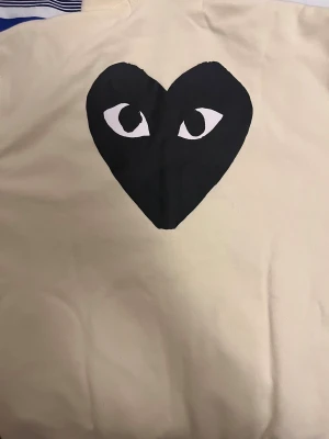 Beige hoodie Comme des Garçons Play - Säljer en beige hoodie från Comme des Garçons Play med det ikoniska svarta hjärtat med ögon tryckt på ryggen och broderat på bröstet. Tröjan har huva och långa ärmar, tillverkad i mjuk bomull. Perfekt för dig som gillar streetwear och vill sticka ut.