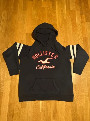 Svart hoodie från Hollister L - Svart hoodie från Hollister med röd och vit broderad logga och text på bröstet. Tröjan har huva med snörning, känguruficka och vita ränder på ärmarna. Tillverkad i mjuk bomullsmix, perfekt för chill dagar.