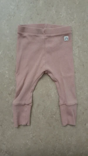 Leggings - Säljer ett par mjuka och stretchiga rosa leggings från Lindex Baby i ribbad bomullskvalitet med elastan. De har breda muddar vid bensluten och en liten tygpatch med regnbågsmotiv framtill. Perfekta för chill dagar hemma.