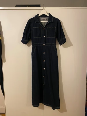 Mörkblå jeansklänning från NA-KD - Supersnygg lång jeansklänning från NA-KD i mörkblå denim med knappar hela vägen framtill, två  bröstfickor och korta ärmar med uppvik. Klassisk krage och markerad midja. Har även två fickor bak. Skitsnygg men tyvärr aldrig använd pga fel storlek.