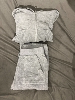 Grå Juicy Couture hoodie och byxor - Säljer en klassisk grå hoodie från Juicy Couture i mjuk velour. Tröjan har huva, långa ärmar, dragkedja med J-detalj och fickor framtill. Dessutom säljer jag juciy byxorna, båda är i storlek S. De har små defekter där metallen är missfärgad, se bilder i slutet. Säljer tröjan för 300kr och byxorna för 300kr, båda för 600kr