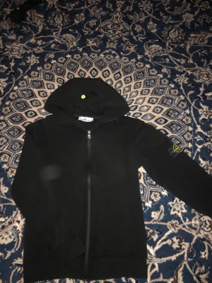 Svart hoodie från Stone Island - Svart hoodie från Stone Island med klassisk logotyp-patch på vänster ärm. Tröjan har huva med gul knappdetalj, hel dragkedja framtill och långa ärmar. Tillverkad i mjuk bomull för skön komfort och stilren look.