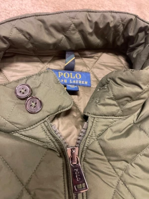 Ralph Lauren jacka  - Quiltad jacka i mellangrön färg från Polo Ralph Lauren. Jackan har ribbade muddar, ståkrage med knappdetaljer och dragkedja framtill. Fodrad insida och två fickor med knappstängning. Perfekt för dig som gillar stilren och klassisk look.