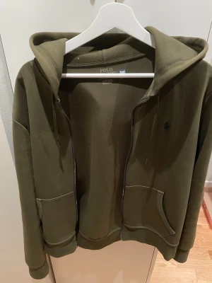 Polo Ralph Lauren zip hoodie - Snygg olivgrön hoodie från Polo Ralph Lauren med dragkedja, två stora fickor framtill och klassisk huva med dragsko. Broderad logga på bröstet. Mjuk bomullsblandning som är skön att ha på sig. Perfekt för en chill och stilren look.