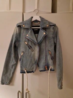 Diesel jeansjacka med dragkedjor - Snygg ljusblå jeansjacka från Diesel med flera dragkedjor i guld och coola detaljer som snörning i nederkant. Jackan har en klassisk bikerkrage, långa ärmar och croppad passform. Perfekt för dig som vill ha en unik denimlook.