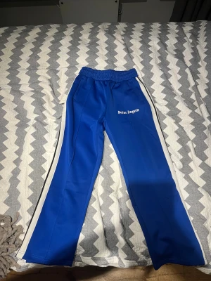 Blå Palm Angels trackpants med vita ränder - Säljer ett par blå trackpants från Palm Angels med vita sidoremsor och logotyp på vänster ben. Byxorna har elastisk midja och dragkedjor vid bensluten. Snygg loose passform och sportig vibe, perfekta för streetwear.