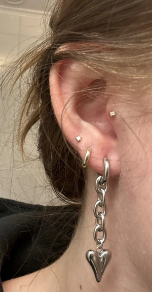 Silvriga örhängen med hjärtberlock - Säljer ett par chunky silvriga örhängen med kedja och hängande hjärtformad berlock. Örhängena har en cool och edgy vibe, perfekta för att lyfta din stil. Materialet är metall med blank finish och hjärtat har en mjukt rundad form.