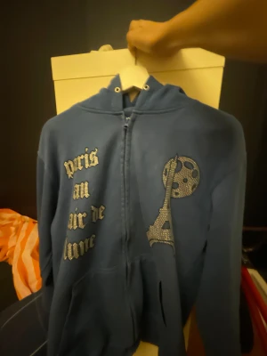 Bling lucid tröja blå - Blå zip-hoodie från BLINGLUCID med glittrande texten 'paris au clair de lune' och strassdekor av Eiffeltornet och måne på bröstet. Tillverkad i 80% bomull och 20% polyester. Klassisk huva och känguruficka framtill.