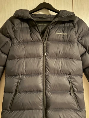 Svart Peak Performance Jacka - Svart dunjacka från Peak Performance med luva och två fickor med dragkedja. Den är i väldigt bra skick utan några synliga fel. 