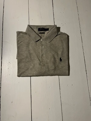 Grå pikétröja från Polo Ralph Lauren - Snygg grå pikétröja från Polo Ralph Lauren med klassisk krage och tre knappar framtill. Tröjan är gjord i mjuk bomull med stretch och har den ikoniska broderade loggan på bröstet. Perfekt för en clean och stilren look.