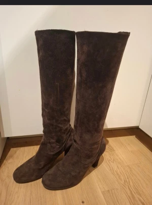 Bruna mockastövlar med klack - Snygga höga stövlar i mörkbrun mocka med rund tå och stabil klack. Stövlarna har en mjuk yta och klassisk form som ger en cool och tidlös vibe. Perfekt för dig som gillar att sticka ut med stilrena boots under höst och vinter.