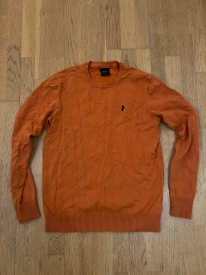 Pullover från Peak Performance - Snygg orange tröja från Peak Performance i storlek M. Tröjan har rund hals, ribbade muddar och ett litet svart broderat logomärke på bröstet. Perfekt för dig som gillar stilrena och enkla plagg med en färgklick.