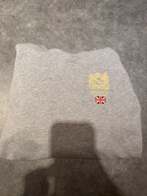 Grå Trenton hoodie från Morris - Grå hoodie från Morris med guldfärgat emblem på bröstet. Tröjan har huva med snörning och är tillverkad i mjuk bomull. Storlek S och sitter som en S. Mycket fint skick och bara använd fåtal gånger.