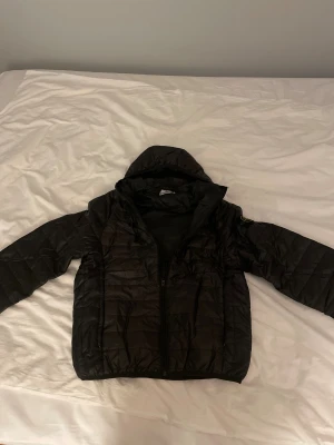 Svart pufferjacka från Stone Island  - Svart pufferjacka från Stone Island med huva och den klassiska patchen på vänster ärm. Jackan har dragkedja framtill och passar perfekt för dig som gillar streetwear och en clean, enkel stil. Den är i storlek M. Jag fick den i present men säljer den eftersom den tyvärr inte passar. Jackan är i princip oanvänd – bara provad några få gånger.