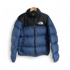 Blå dunjacka från The North Face - Säljer en fet blå dunjacka från The North Face med svart axelparti och logga på bröstet. Jackan har hög krage, två sidofickor med dragkedja och en rejäl puffer-look. Perfekt för kalla dagar och riktigt snygg streetstil.